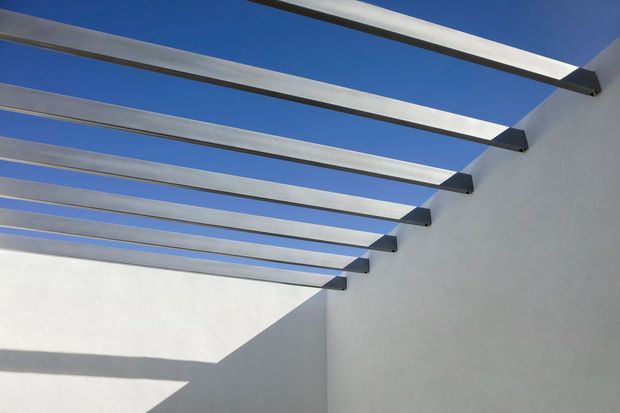 Minimalist architectural detail of an open-air pergola or covered terrace, with evenly spaced horizontal metal beams projecting from smooth white plaster walls. Strong sunlight creates crisp geometric shadows, with vivid blue sky visible through the open roof.