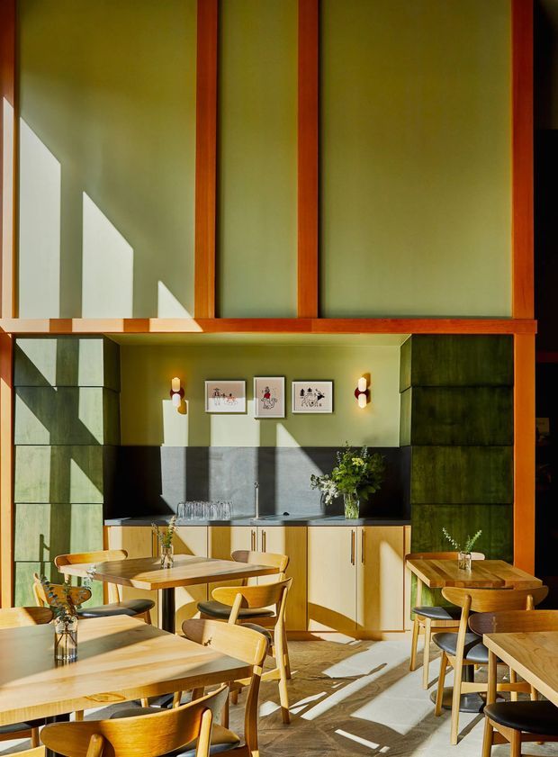 A sunlit contemporary cafe or dining space with a dramatic double-height volume, large timber-framed glazed panels, olive-green walls, and strong geometric shadows. The room contains multiple light timber dining tables and chairs, a compact service counter with pale cabinetry and a dark splashback, framed artwork, wall sconces, and plants. Warm natural light and timber furnishings create an inviting, refined atmosphere.