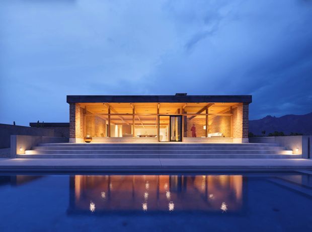 A modern single-storey pavilion-like residence photographed at dusk from across a reflecting swimming pool. The building has a strong flat roof, extensive floor-to-ceiling glazing, warm timber-lined interior surfaces, and a broad stone or concrete terrace with wide stepped access to the pool. Integrated low-level lighting emphasizes the horizontal composition, while dark mountain silhouettes form the distant backdrop.