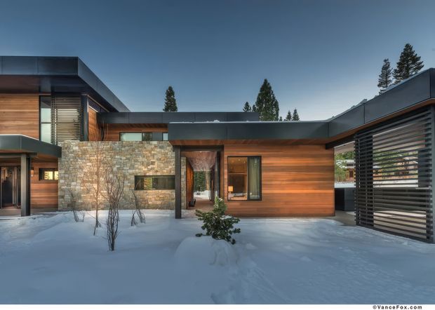 Exterior view of a contemporary low-rise residence in a snowy setting. The home features horizontal warm timber cladding, dark metal roof planes and window frames, a prominent natural stone wall, extensive horizontal louvres/screens, and a sheltered entry courtyard. Tall evergreen trees and deep snow surround the building.