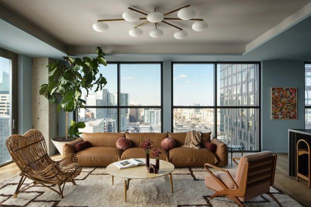 A stylish high-rise living room with expansive floor-to-ceiling corner glazing overlooking a dense urban skyline. The room features a warm brown leather sofa, sculptural lounge chairs, a round coffee table, a textured neutral rug, a large indoor tree, abstract artwork, and a distinctive multi-globe ceiling light. Dark window frames and muted blue-grey walls create a refined contemporary setting.