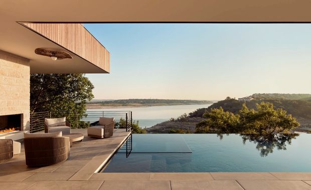 A contemporary waterfront terrace with an infinity pool overlooking a calm bay or lake. The space features a cantilevered roof with timber soffit detailing, pale stone or masonry walls, large-format terrace paving, outdoor lounge furniture, a metal balustrade, and a prominent tree-covered shoreline beyond.