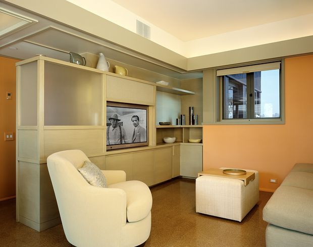 Warm mid-century-inspired living room with built-in timber cabinetry, open display shelving, an integrated television, upholstered armchair and ottoman, and a large sofa. The room has an orange accent wall, neutral carpet, recessed ceiling detailing, and a window overlooking neighboring buildings.