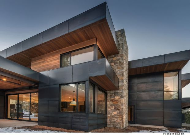 Exterior architectural photograph of a contemporary multi-level home with dark metal cladding, extensive warm timber soffits, floor-to-ceiling glazing, and a prominent rough-stacked natural stone chimney or wall element. Strong horizontal roof overhangs and projecting volumes define the modern composition. Snow is visible at ground level, but the focus is the building facade.