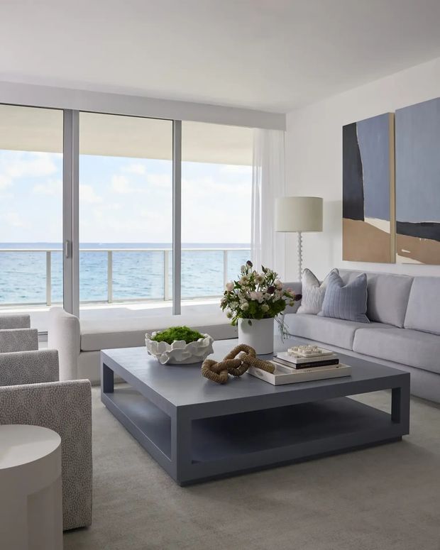 Bright, refined living room with a large grey sectional sofa, matching textured armchairs, a dark square coffee table, pale rug, sculptural decor, floral arrangement, abstract wall art, and a floor lamp. Full-height glazing opens to a balcony with expansive ocean views, creating an airy coastal atmosphere.