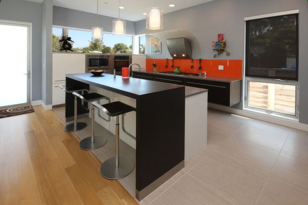 Contemporary kitchen featuring a large dark island with integrated seating, pale cabinetry, grey walls, pendant lighting, and a vivid orange glass splashback. Timber flooring transitions to light tile near the kitchen work area, with generous windows and a glazed door providing natural light.