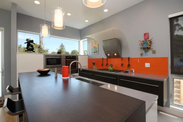 Contemporary kitchen with a large dark island bench, black fixed cabinetry, stainless-steel sink and appliances, and a vivid orange splashback. Large corner windows provide abundant daylight, while cylindrical pendant lights hang above the island. The room includes playful wall artwork and a sculptural rangehood enclosure.