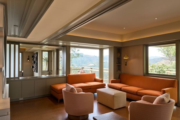 A warm, refined living room with expansive windows overlooking a hilly landscape. The space features a large orange-brown sectional sofa, matching lounge chairs, a pale upholstered ottoman, dark timber cabinetry, integrated shelving, recessed ceiling lighting, and a mirrored or reflective wall section. Floor-to-ceiling glazing and generous seating create an inviting, visually connected indoor-outdoor setting.