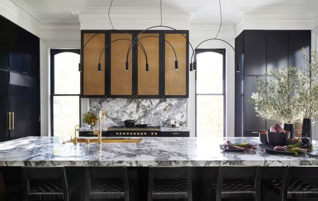 A sophisticated kitchen with deep navy-black cabinetry, a large white marble island and matching marble splashback, a professional range cooker, and a dramatic woven rattan feature canopy above the cooking zone. Black pendant lights hang over the island, which includes seating for several people. Tall windows and ornate white ceiling cornices bring in natural light and add a refined traditional character.