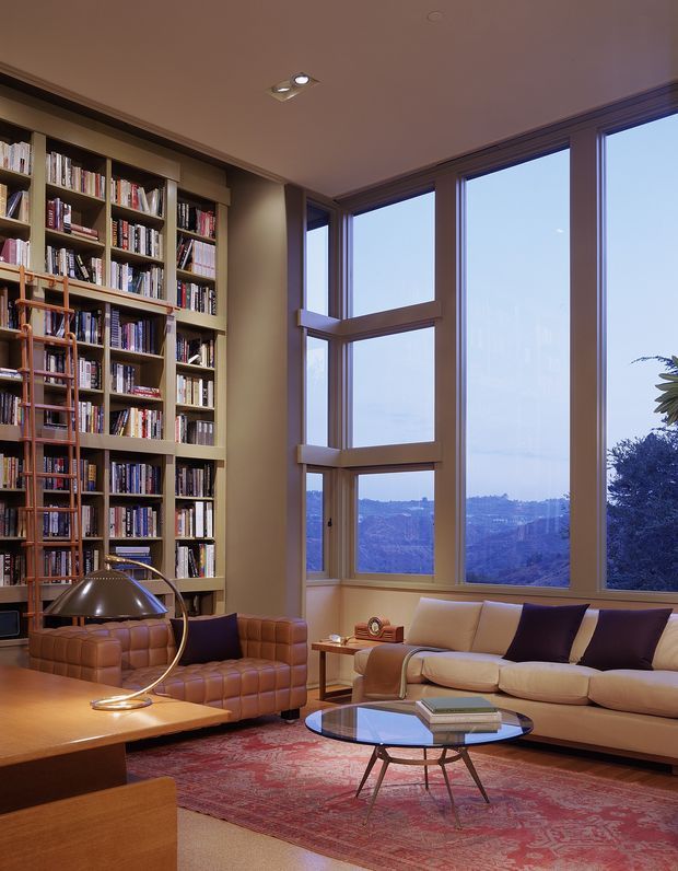 A warm, refined living room with a dramatic floor-to-ceiling corner window overlooking a distant landscape. A full-height built-in timber bookcase fills the left wall, while a cognac leather armchair and matching ottoman sit beside a cream sofa. A low round glass coffee table, sculptural floor lamp, patterned red rug, and timber furnishings complete the sophisticated interior.