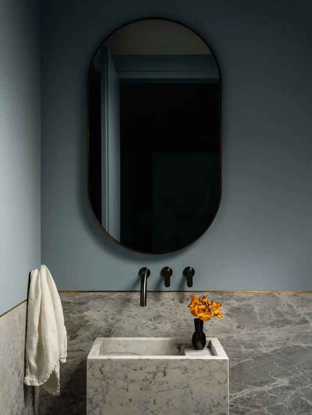 A moody, refined bathroom vanity featuring a large vertically oriented rounded-arch mirror with a dark frame, dark wall paint, a wall-mounted black tap and controls, and a sculptural rectangular marble vessel basin. Grey veined stone continues across the backsplash and side walls, with a slim warm-toned trim at the transition. A white towel and small vase of dried orange flowers add restrained accents.