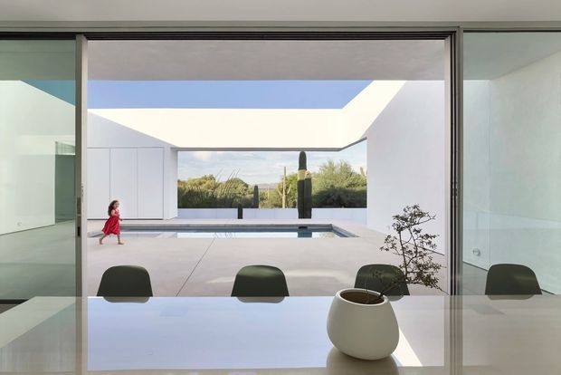A contemporary dining area viewed through fully glazed sliding doors toward a minimalist courtyard pool. The interior has a glossy white dining table with dark chairs and a simple ceramic planter, while the courtyard features pale paving, white walls, a linear pool, and a large open roof aperture framing the sky and landscape. A child is visible outdoors, but the architecture and strong indoor–outdoor connection dominate the composition.