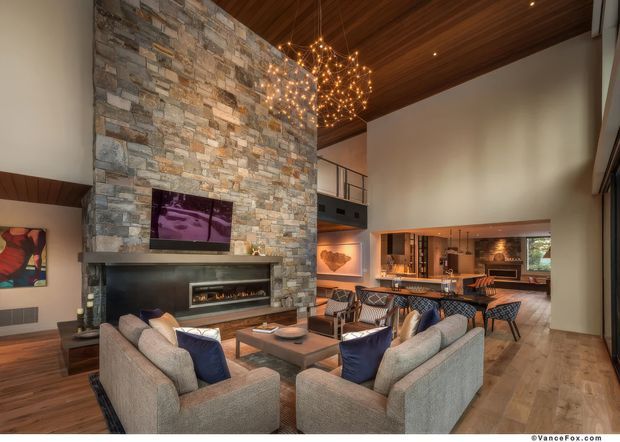 Spacious, high-ceilinged open-plan living room with a dramatic stacked-stone fireplace wall, built-in linear fire, and wall-mounted television. The room features warm timber flooring and a timber-clad ceiling, contemporary sofas and armchairs arranged around a coffee table, and a sculptural starburst pendant. An open dining and kitchen area is visible beyond, with a mezzanine-style railing and large glazing contributing to the airy lodge-like setting.