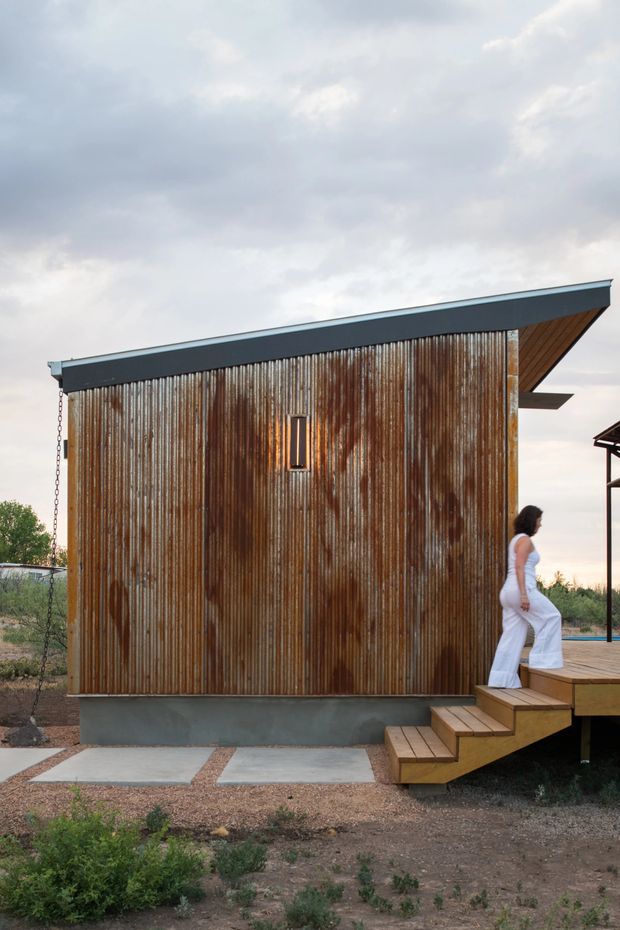 Exterior view of a compact modern building with a strongly sloped roof and corrugated weathered metal wall cladding. A small vertical window punctuates the facade, while a timber stair leads up to an elevated timber deck. The structure sits in a sparse landscaped setting under an overcast sky.