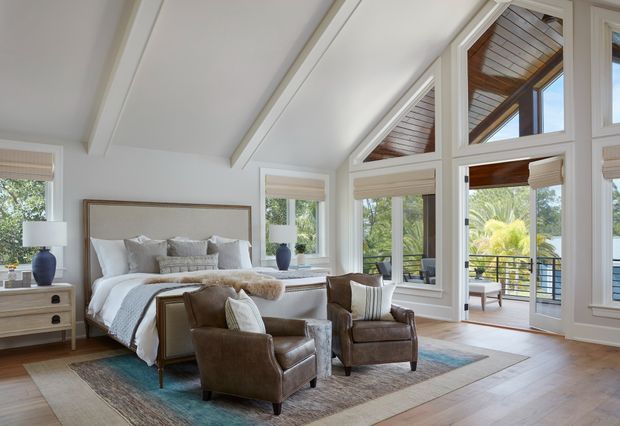 Bright, refined bedroom with a vaulted ceiling and large triangular clerestory windows overlooking a leafy outdoor setting. The room features a substantial upholstered bed, layered neutral bedding, a pair of leather armchairs, bedside tables, area rugs, and warm timber flooring. Glazed doors open to a balcony or terrace, while the vaulted ceiling continues into a timber-lined exterior or transition space.