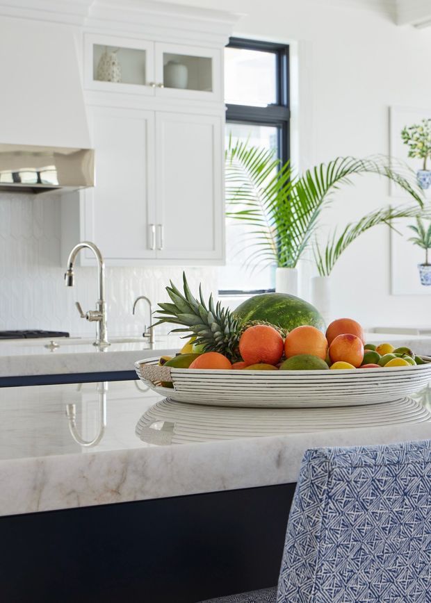 Bright, refined kitchen interior with white Shaker-style cabinetry, glazed upper cupboards, dark-framed windows, a white geometric tile splashback, and a large pale marble island with a dark base. A decorative bowl of fruit sits on the island, with tropical plants and blue patterned upholstery adding colour and texture.