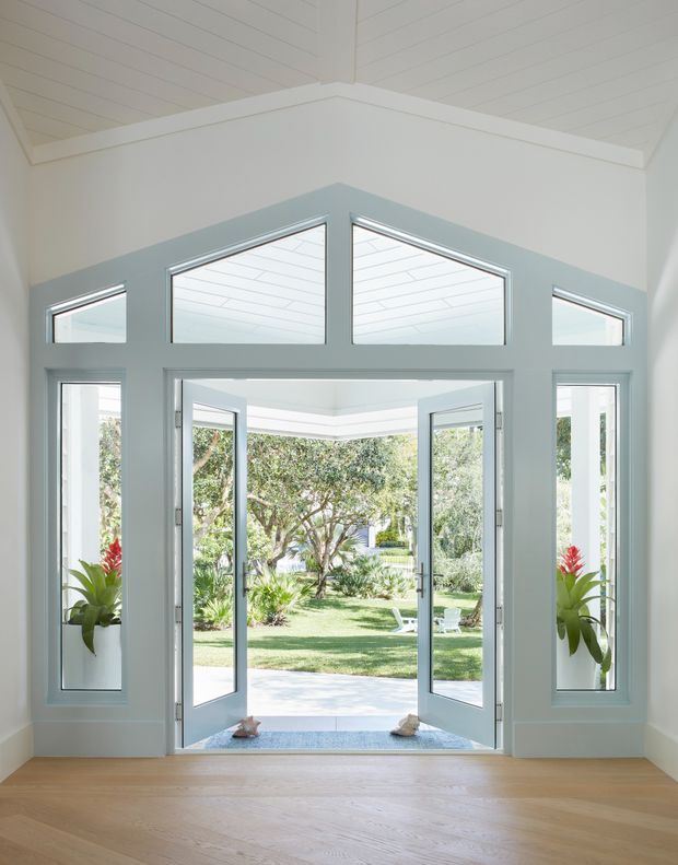Bright, airy entryway with pale blue-grey and white trim surrounding a pair of open glazed doors, narrow sidelights, and triangular clerestory windows. The room has light timber flooring and a white painted timber-lined ceiling, opening directly onto a landscaped garden.