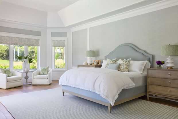 Bright, elegant bedroom with a large upholstered bed, pale blue-grey curved headboard, white bedding, matching bedside tables, and a classic wooden chest of drawers. The room features light grey textured wallcovering, white painted trim and ceiling panelling, tall windows with patterned Roman blinds, and a soft striped area rug. A small sitting area with two armchairs occupies the window bay.