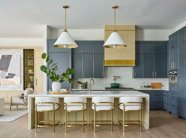 A refined open-plan kitchen with deep blue-grey cabinetry, brass hardware, a pale stone island benchtop, and a large timber-and-brass rangehood surround. Four upholstered bar stools line the island beneath two white and brass pendant lights. A timber-floored living area, shelving, artwork, and greenery are visible beyond the kitchen.