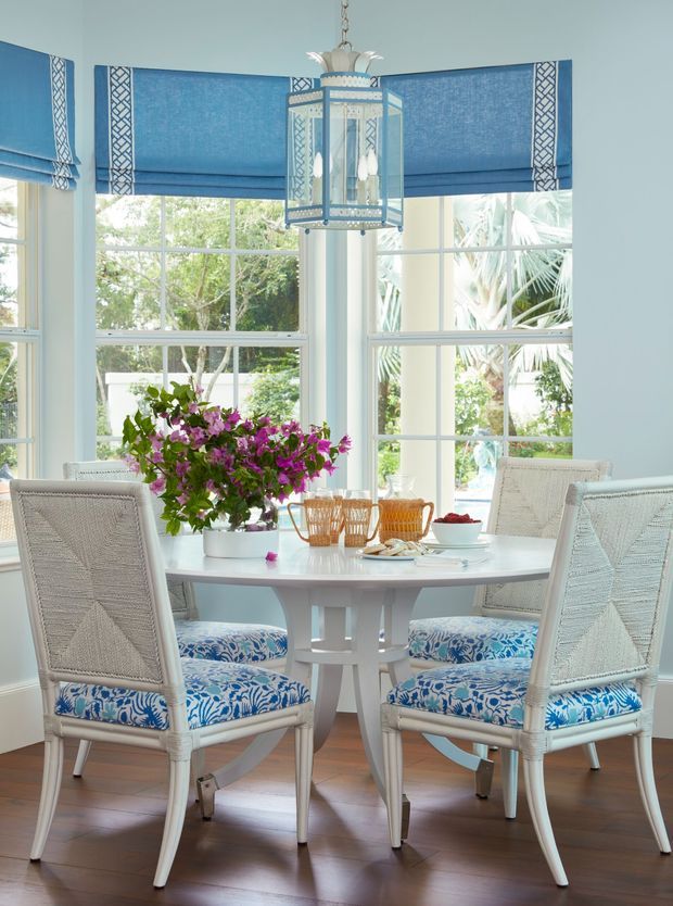 Bright, elegant dining nook with a round white dining table surrounded by woven white chairs upholstered in blue-and-white botanical fabric. Large multi-pane bay windows with blue Roman blinds overlook lush greenery, while a decorative blue-and-white pendant lantern hangs above. Dark timber flooring and abundant natural light create a fresh coastal feel.