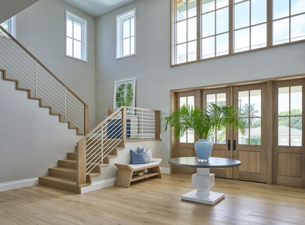 A bright, double-height residential entry hall with a timber staircase, horizontal metal balustrades, large grid-pane windows, and glazed timber double doors. Light timber flooring, pale walls, a small bench, round table, and indoor palm create an airy, refined foyer.