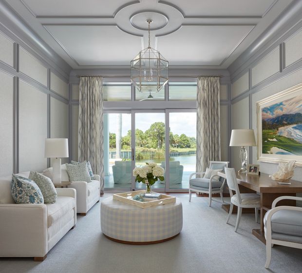 Elegant, symmetrical sitting room with grey paneled walls, a decorative coffered ceiling, large glazed doors opening to a leafy water view, patterned curtains, upholstered sofas and armchairs, a rounded ottoman coffee table, and a small writing desk. A geometric pendant light and framed landscape artwork add refined decorative detail.