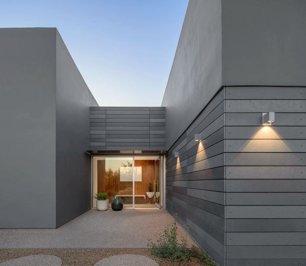 A minimalist modern residential entry courtyard viewed from outside, framed by tall smooth grey rendered walls and a dark horizontal-panel facade. A glazed entrance door reveals a warm timber-lined interior, with subtle wall-mounted lighting illuminating the pathway and sparse potted plants near the doorway.