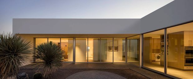 A low-slung contemporary single-storey residence arranged around a landscaped courtyard at dusk. The home has crisp flat roof planes, extensive floor-to-ceiling glazing with dark slender frames, warm interior lighting, and a visible open-plan kitchen through the right-hand glass wall. Spiky ornamental planting and gravel landscaping define the foreground.