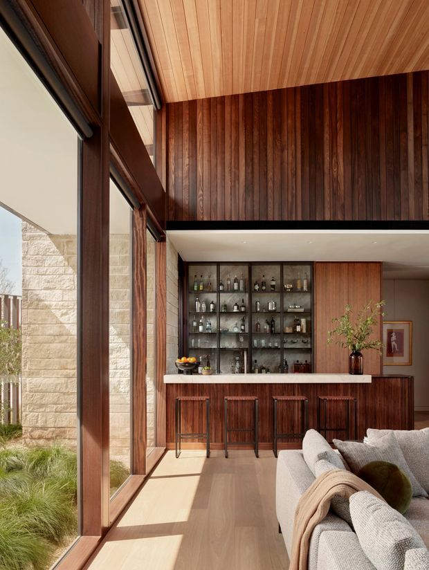 A refined home bar and lounge area with extensive dark timber wall panelling, a matching slatted bar front, glass-fronted bottle cabinetry, a pale stone or solid-surface countertop, and four bar stools. Floor-to-ceiling glazing opens toward a landscaped courtyard, while a large upholstered sofa occupies the foreground. The ceiling continues the warm timber lining, creating a cohesive, richly detailed interior with strong natural light.