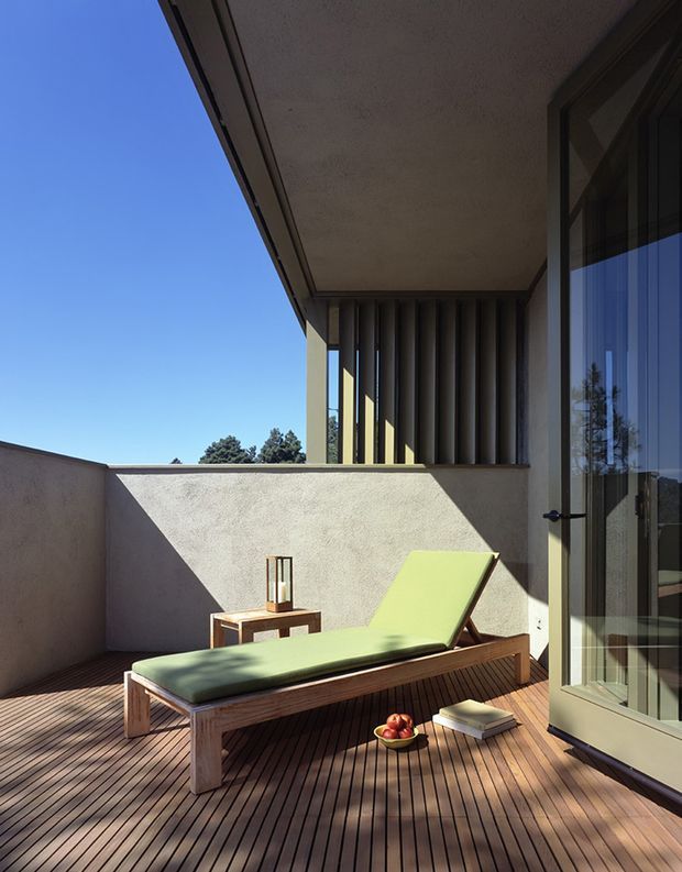 Sunlit elevated timber deck terrace with a green upholstered chaise lounge, small side table, lantern, and books. The terrace is enclosed by rendered walls and a deep roof overhang, with dark vertical screening fins and large glazed doors creating a private outdoor retreat. The architecture has clean modern lines and strong geometric shadows.
