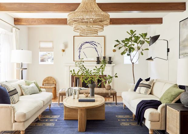 Bright, inviting living room with two cream sofas arranged around a rounded timber coffee table, a blue patterned rug, indoor plants, a fireplace, woven pendant light, and exposed timber ceiling beams. The room combines warm natural textures with layered neutral furnishings and dark blue accents.