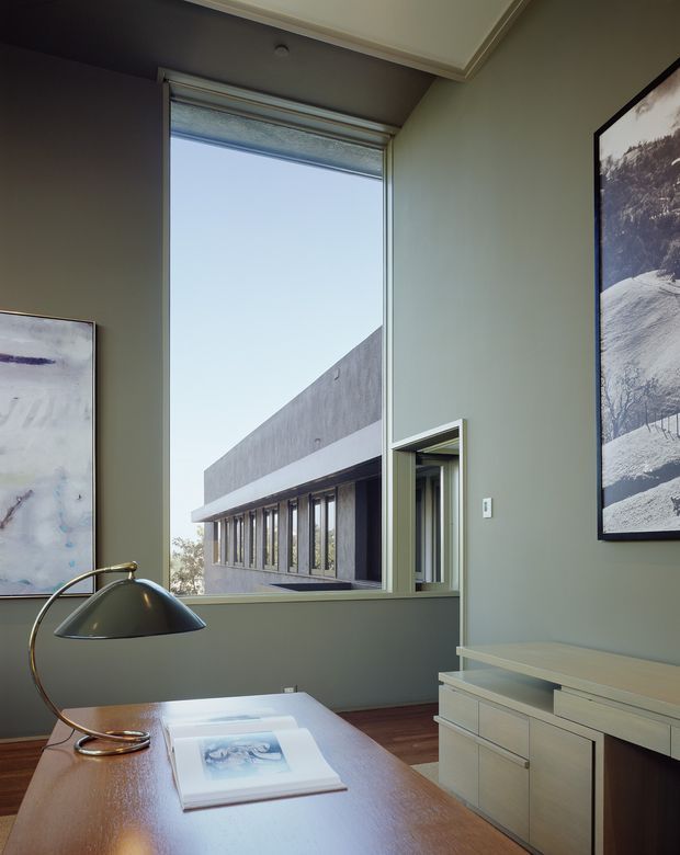 A refined home office with muted sage-grey walls, a large vertical window, and built-in low cabinetry. The room contains a wood desk with a sculptural brass-and-black desk lamp and an art book, framed artwork, timber flooring, and a partial view of a modern neighboring building through the window. The space has a calm, contemporary feel with tailored trim and restrained furnishings.