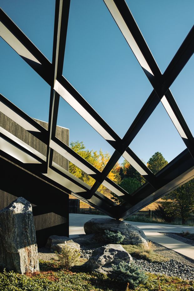 Exterior architectural view of a dramatic black steel diagonal structural frame or canopy extending over a landscaped courtyard. The angular exposed beams contrast with a clear blue sky, dark timber-clad walls, large natural boulders, gravel, and low planting. The image emphasises sculptural structure, strong geometric lines, and an integration of architecture with a carefully designed landscape.