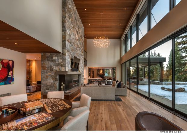 Spacious contemporary living room with a dramatic double-height volume, full-height glazing, exposed warm timber ceiling, natural stone fireplace wall, timber flooring, and layered lounge and dining seating. A snow-covered outdoor setting is visible through the glazing, while a sculptural chandelier hangs over the room.