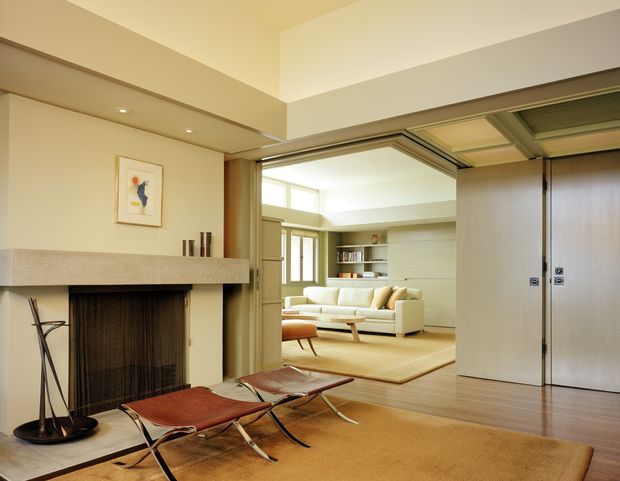 View from a refined living room into an adjoining lounge through large sliding timber-framed doors. The foreground features a broad fireplace with a simple stone mantel, leather sling chairs, a low timber table, and warm wood flooring partly covered by a neutral rug. The connected lounge has a pale sofa, built-in shelving, light cabinetry, and abundant clerestory-style glazing. The palette is warm and understated, with clean-lined mid-century furnishings and an open, airy composition.