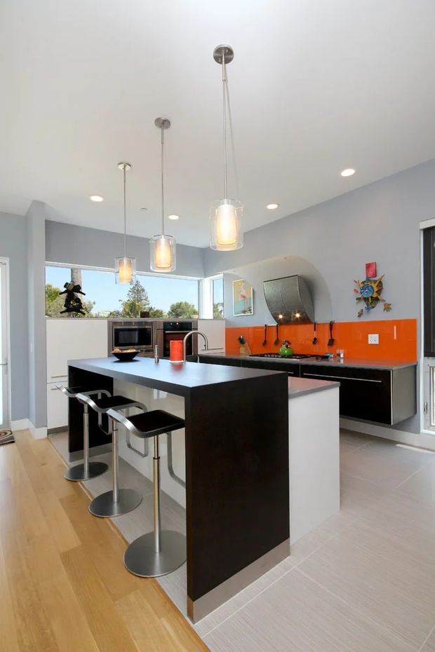 Contemporary open-plan kitchen dominated by a large black island with a white waterfall-style side panel and three modern bar stools. The room has light grey walls, a bright orange glass or glossy splashback, dark cabinetry, pale tiled flooring transitioning to timber flooring, stainless-steel appliances, recessed ceiling lights, and three pendant lights. Large windows provide abundant daylight and views of greenery.