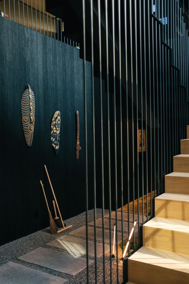 A dramatic interior stairway with pale timber stair treads beside a tall black metal vertical-rod balustrade. The adjacent dark textured wall displays carved decorative masks, while large stone stepping slabs and dark gravel create a tactile transition at floor level. The space is softly lit with strong contrast and a refined, moody atmosphere.