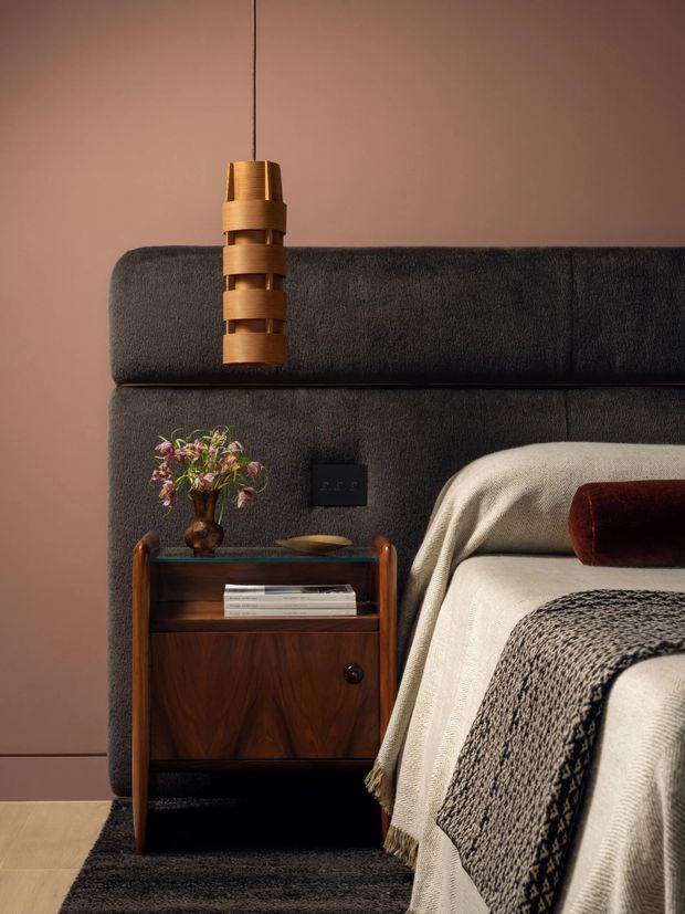 A close-up bedroom vignette featuring a dark upholstered bed with a tall padded headboard, cream bedding, a textured charcoal throw, and a warm walnut bedside cabinet. A sculptural timber pendant hangs above the bedside table against a muted terracotta wall, creating a warm, refined and tactile atmosphere.