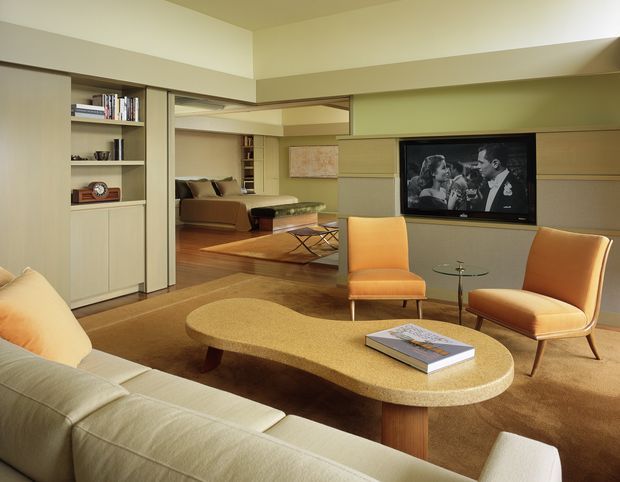 Warm, mid-century-inspired living room with a cream sectional sofa, sculptural oval coffee table, two orange upholstered lounge chairs, and a wall-mounted television. Built-in pale wood shelving and cabinetry frame a wide opening to a secondary sitting area, with timber flooring and muted beige finishes throughout.