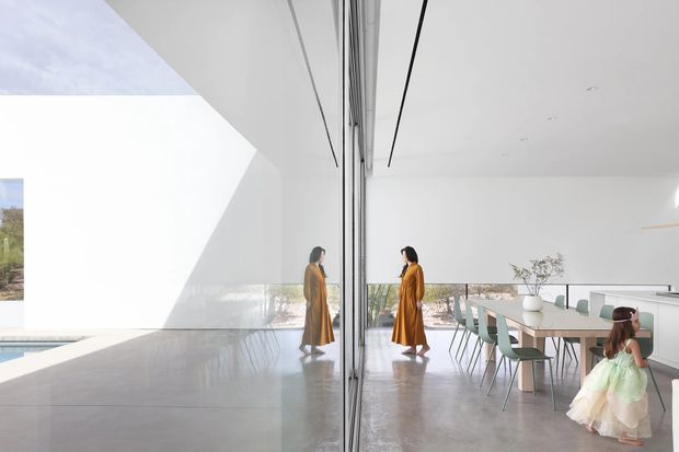 Bright, minimalist contemporary dining area with a long timber dining table, pale upholstered chairs, and expansive floor-to-ceiling glazing. A mirrored glass partition reflects the dining space and figures, while sculptural white walls, polished concrete flooring, and a strong geometric opening create a gallery-like interior. A glimpse of the pool and landscape is visible outside.