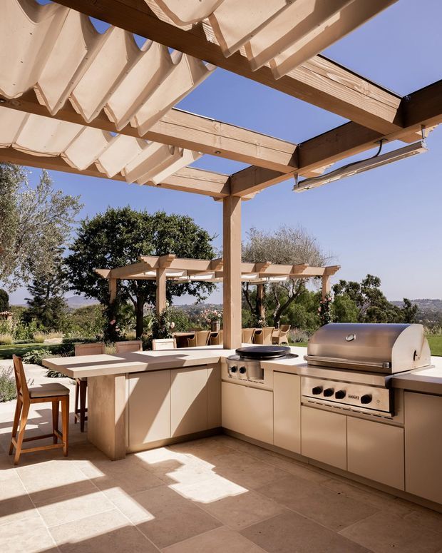 A bright, spacious outdoor kitchen and entertaining area beneath a timber pergola with retractable fabric shade panels. The built-in kitchen includes beige cabinetry, stone-look benchtops, a gas barbecue and cooktop, with a dining counter and stools. Pale tiled flooring, timber posts, landscaped grounds and distant hills create a warm, resort-like setting.