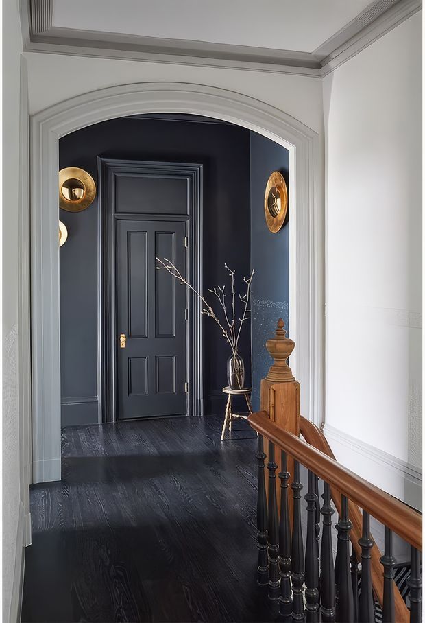 A refined residential hallway or landing viewed from a staircase, featuring a dramatic dark navy-black paneled door and wall set within a large curved archway. The space has dark timber flooring, white decorative wall panelling and ceiling mouldings, brass circular wall accents, and a timber stair handrail with black turned balusters. A small wooden pedestal and vase with branches add understated decor.