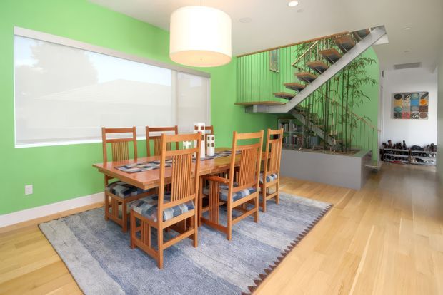 Bright open-plan dining area with a solid timber dining table and matching high-back chairs on a large grey rug. A vivid green painted wall, large window with roller blind, pale timber flooring, and a sculptural open-riser staircase with green vertical balustrade and integrated planter define the space. Artwork and a shoe-storage area are visible in the background.