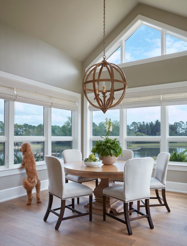 Bright dining area with a round wooden table and upholstered bar-height dining chairs, set within a window-filled corner overlooking water and trees. The room has a vaulted ceiling, large white-framed windows with woven shades, light timber flooring, and a prominent bronze-toned spherical pendant light. A potted plant and dog are also visible.