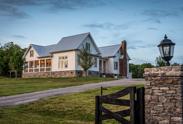 Exterior view of a large light-coloured farmhouse-style residence with multiple pitched gable roofs, standing-seam metal roofing, white weatherboard cladding, stone masonry at the base and chimney, and a covered front porch. A gravel driveway, lawn, timber gate, and stone entrance pillar with lantern frame the property.