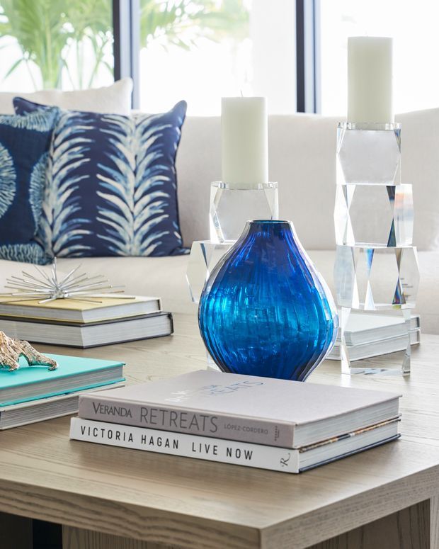 A bright, styled living room vignette featuring a pale timber coffee table layered with design books, a vivid cobalt-blue glass vase, geometric crystal candleholders with white pillar candles, and a navy patterned cushion on a light sofa. Large windows bring in abundant natural light and greenery is visible beyond.