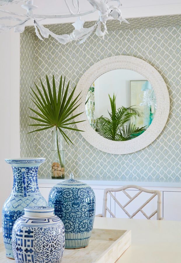 A bright, highly styled dining area with pale green and white geometric patterned wallpaper, white wall panelling, a large round textured white-framed mirror, tropical palm foliage, blue-and-white ceramic vessels, and a light rattan dining chair. A sculptural white chandelier hangs overhead, creating a fresh coastal and Hamptons-inspired look.