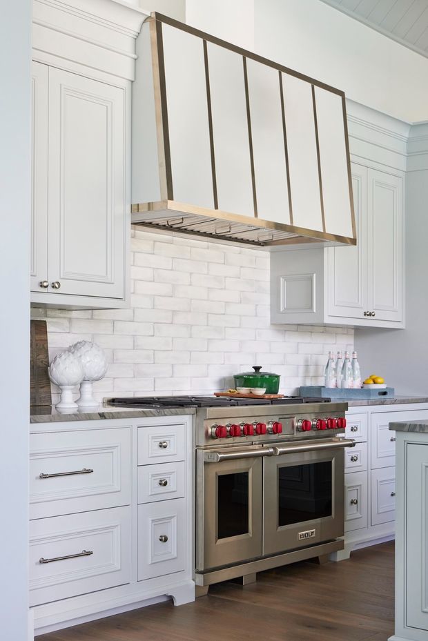 Bright, refined kitchen featuring white raised-panel cabinetry, a large stainless steel range cooker, marble-look countertops, white subway tile backsplash, and a custom metal-and-glass range hood surround. Warm timber flooring and polished metal hardware add contrast.