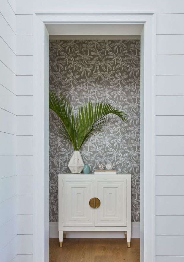 A narrow decorative alcove framed by white moulding and horizontal white wall panelling. The alcove has grey botanical patterned wallpaper, a white paneled console cabinet with brass circular pulls, and a tall leafy palm arrangement in a faceted vase, with warm timber flooring below.