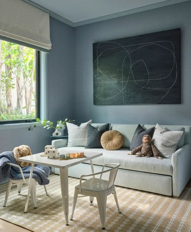 A bright, stylish living room with a pale upholstered sofa, layered grey and cream cushions, a round tan accent cushion, small white coffee table, metal dining-style chairs, patterned rug, indoor plants, and a large abstract dark artwork. The room has blue-grey walls, white trim, a large window with a light Roman blind, and a calm contemporary family-friendly feel.